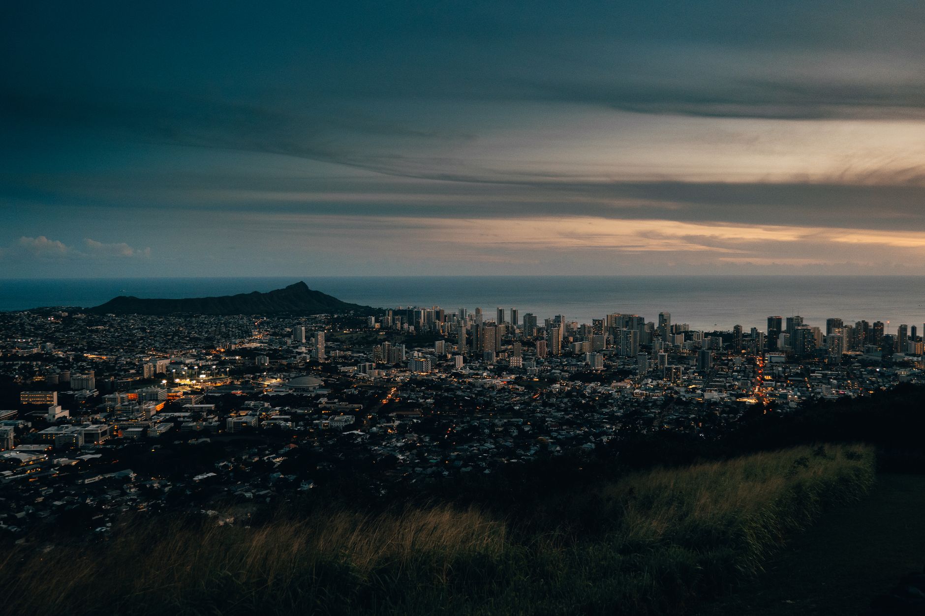 Honolulu skyline at dusk with Diamond Head crater silhouetted against the coastline, the dense urban core of Oahu where shelter and disaster-response sites stand up