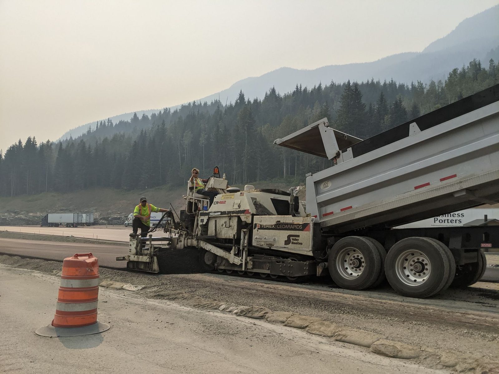 A paving crew and dump truck laying new highway lanes with forested mountains behind